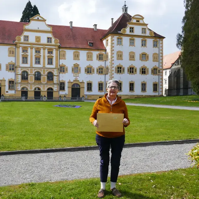2021.4.15__Abgabe_Kleindenkmale__2_.webp Elisabeth Lohrer, Ehrenamtliche Referentin für Geschichte und Dorfentwicklung der Gemeinde Sipplingen steht vor Schloss Salem und hölt einen Briefumschlag in der hand