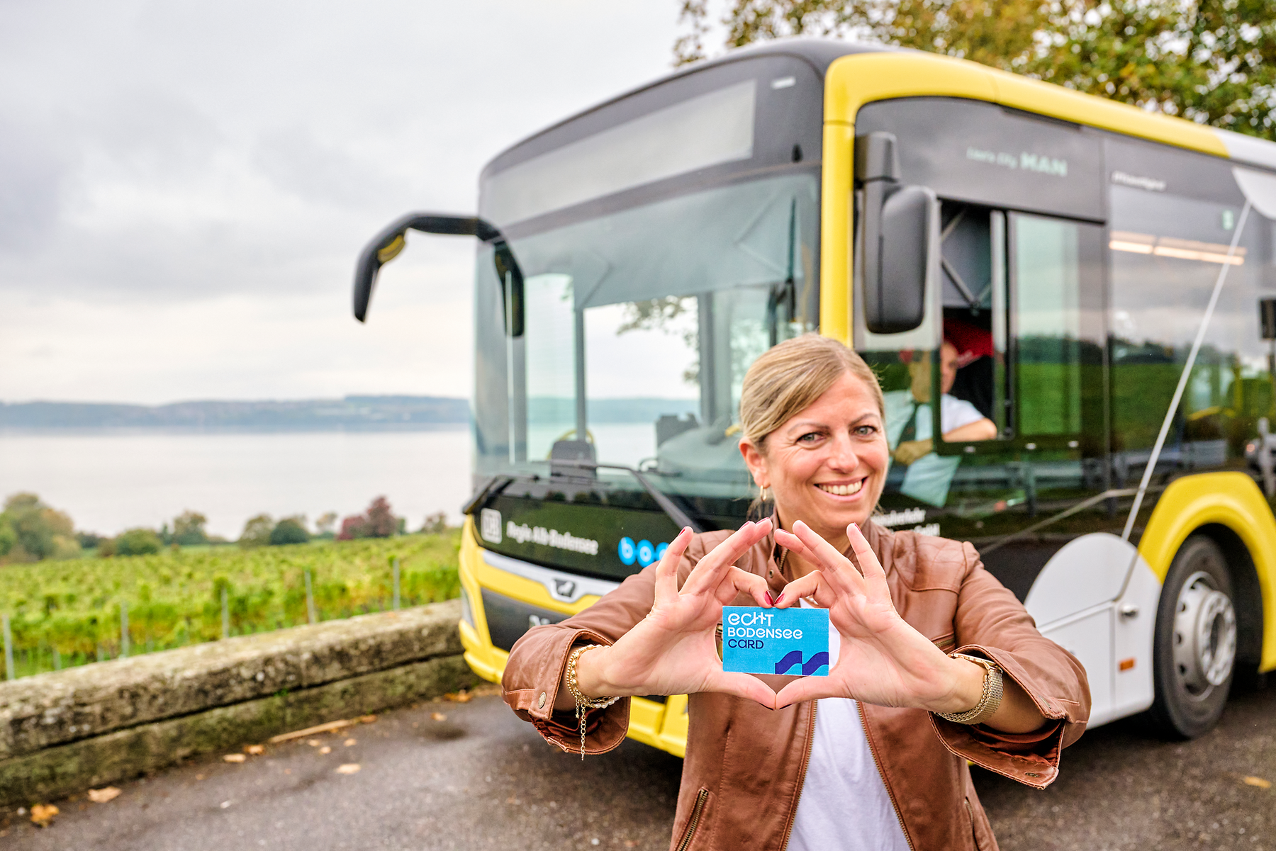 Eine Frau hält ihre Gästekarte vor sich. Im Hintergrund ist ein Bus und der Bodensee.