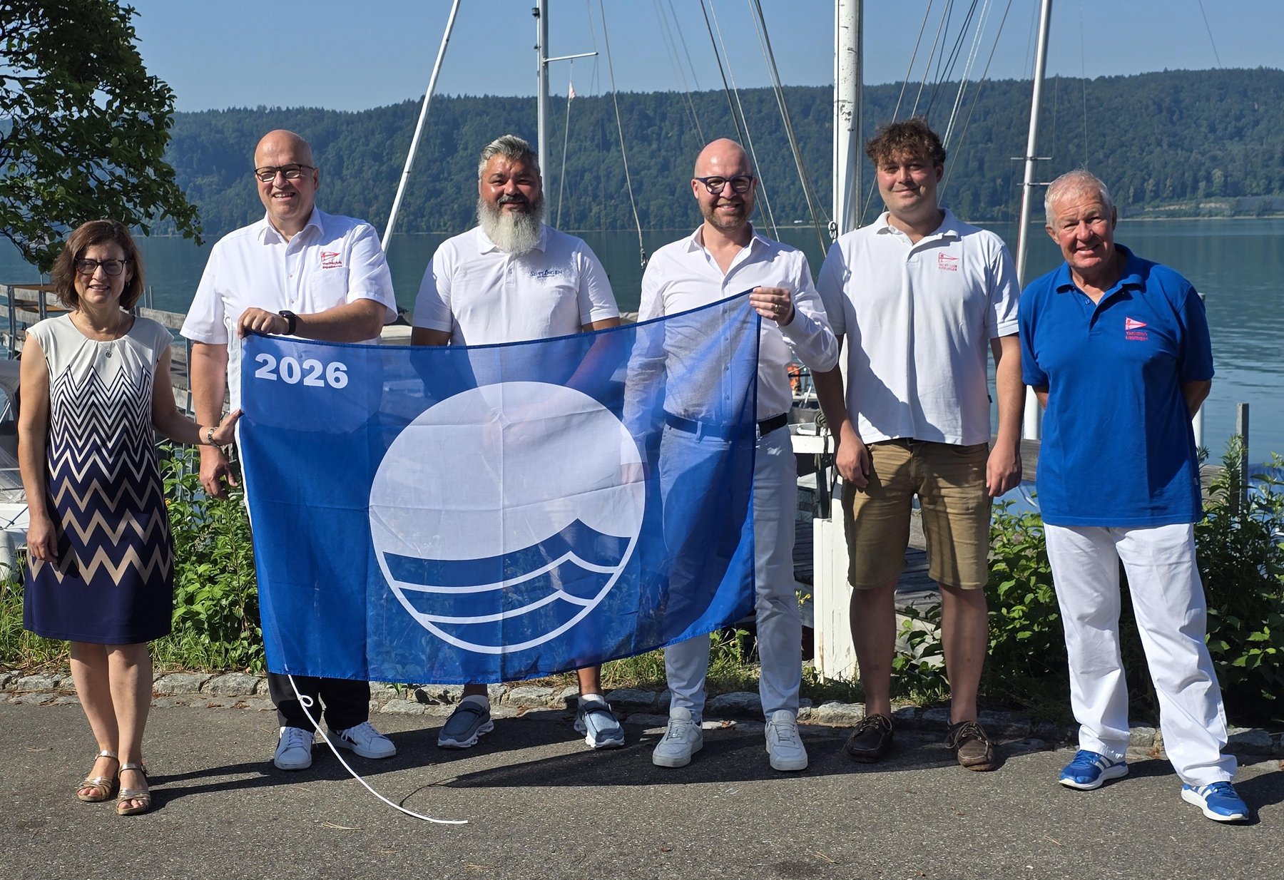Eine Frau und 5 Männer halten die blaue Flage vor sich . Im Hintergrund der Bodensee.